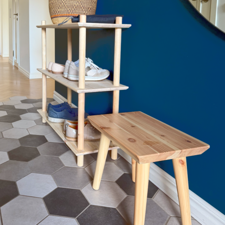 Lifestyleaufnahme mit dem dreistöckigen Air Regal aus unbehandeltem Kiefernholz, das mehrere Paar Schuhe trägt, platziert neben dem Air Hocker – vor einer dunkelblauen Wand und auf einem gemusterten Boden im Eingangsbereich.