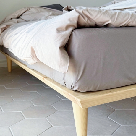 Nahaufnahme des konisch zulaufenden Holzbeins und der Rahmenecke eines Air Bettgestells 140x200 cm aus unbehandeltem, hellem Kiefernholz mit Bettwäsche auf einem sechseckigen Fliesenboden.