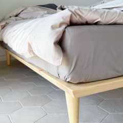 Gros plan du pied en bois effilé et de l’angle du cadre de lit Air 160 × 200 cm en pin clair non traité, avec literie, posé sur un sol carrelé à motifs hexagonaux.
