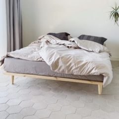 Cadre de lit Air 140 × 200 cm en pin clair non traité, avec une literie gris clair et beige, placé sur un sol carrelé hexagonal clair dans une chambre minimaliste.