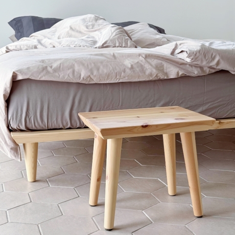 Ein Air Hocker aus unbehandeltem, hellem Kiefernholz steht neben einem Air Bettgestell 160x200 cm mit grauer und heller Bettwäsche auf einem sechseckigen Fliesenboden.