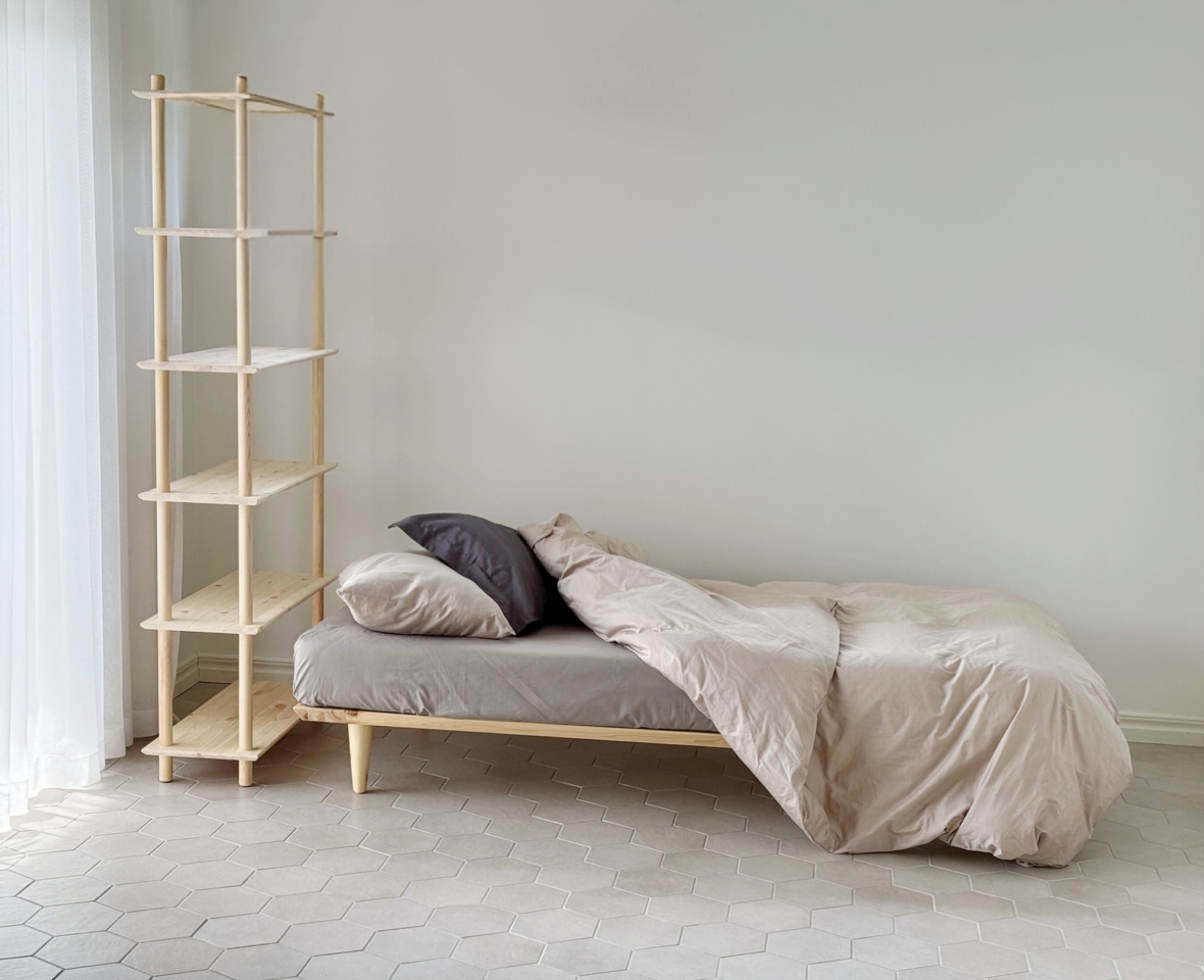 Ein Air Bettgestell 90x200 aus unbehandeltem hellem Kiefernholz mit neutraler Bettwäsche steht neben einer hohen modularen Air Regal-Einheit in einem hellen, minimalistischen Raum.