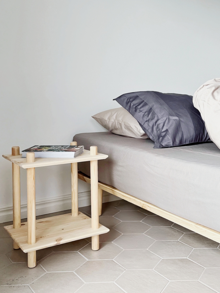 Lifestyle-Aufnahme des Rohe zweistufigen Regals aus rohem Kiefernholz, das als Nachttisch neben einem Bett mit Kissen dient und einen kleinen Stapel Bücher hält.