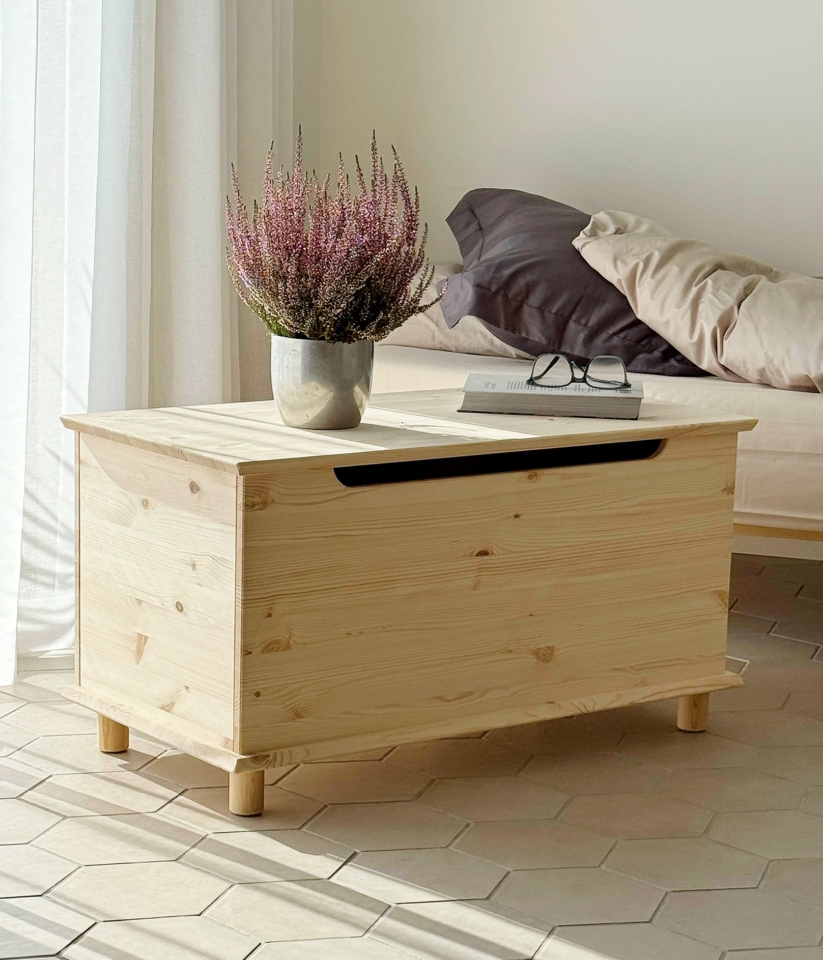 An untreated light pine Air chest with a potted plant and eyeglasses on top, positioned next to an Air bed frame with neutral bedding, on a tiled floor.