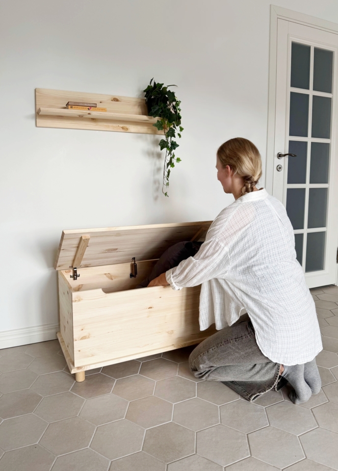 A young woman is kneeling beside an open untreated light pine Air chest, placing or retrieving items. An Air wall shelf with a plant is mounted above on a white wall.