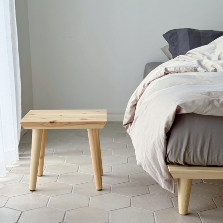 An untreated light pine Air stool stands on a light hexagonal tiled floor next to a Air bed with neutral bedding, seen from a lower angle.