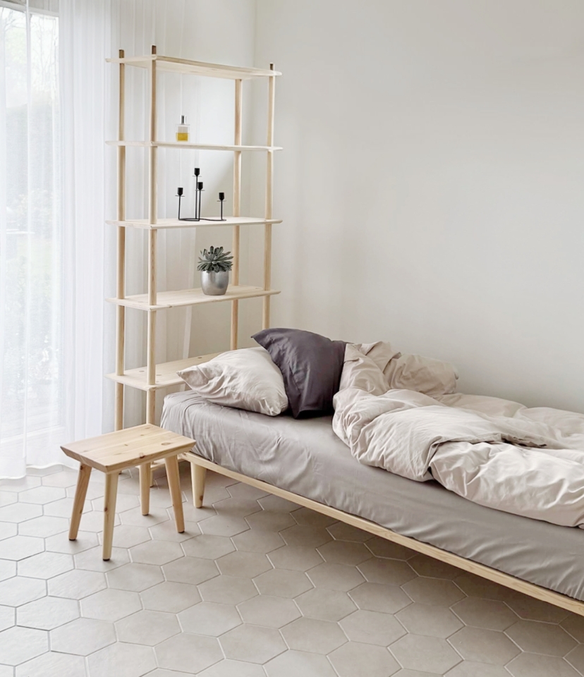 A light-filled bedroom with a simple wooden bed, an untreated pine Air stool, and a tall wooden Air shelf unit, all part of a minimalist aesthetic on a tiled floor.