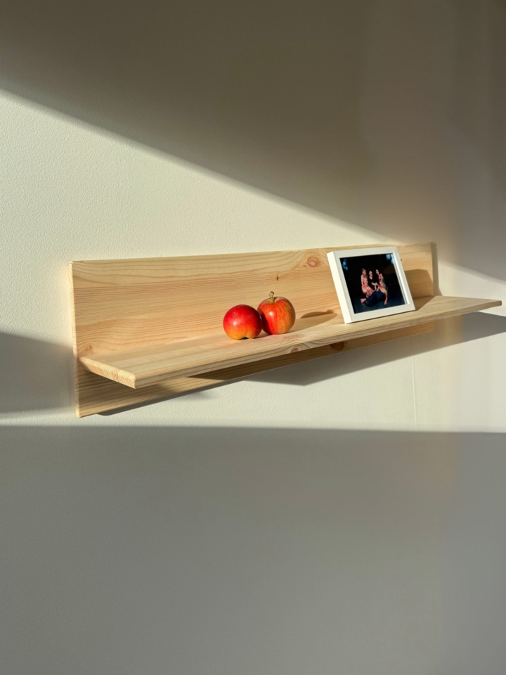 Lifestyle image of the Rohe Air wall shelf, made of raw pine, mounted on a white wall, displaying a photo frame and two apples, illuminated by natural sunlight.