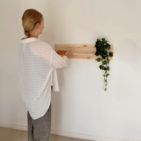A young woman placing a photo frame on the raw pine ROHE Air Wall Shelf, which is styled with a hanging green plant to demonstrate simple and natural accessorizing.