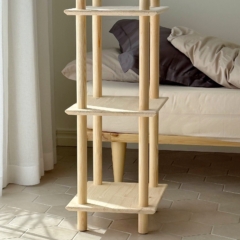 Lifestyle shot of the raw pine Air Shelf narrow three-tiered modular unit in a bedroom corner, next to a bed with pillows, illuminated by natural light.