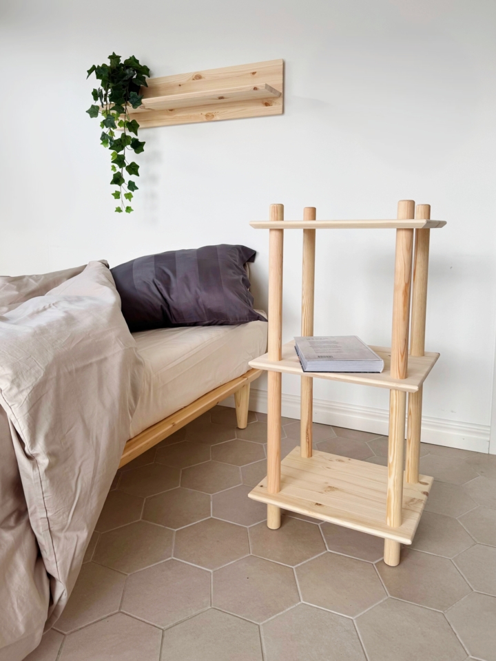 Lifestyle shot of the raw pine Air Shelf narrow three-tiered modular unit next to a bed, holding a book, with a matching wall shelf above it, styled with a trailing ivy plant.