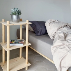 Lifestyle shot of the raw pine Air Shelf narrow three-tiered modular unit next to a bed with grey and beige bedding, holding a succulent plant in a silver pot and a book.
