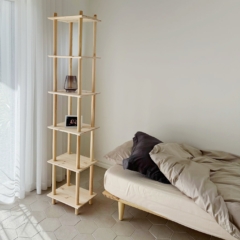 A tall untreated light pine Air modular shelf with a vase and photo frame stands next to an Air bed frame with neutral bedding, in a minimalist room with natural light.