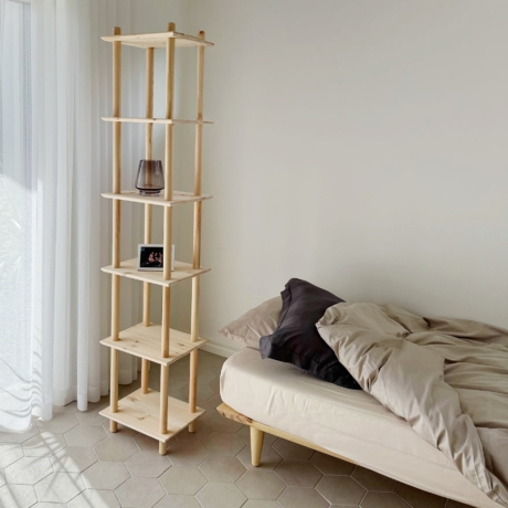 A tall untreated light pine Air modular shelf with a vase and photo frame stands next to an Air bed frame with neutral bedding, in a minimalist room with natural light.