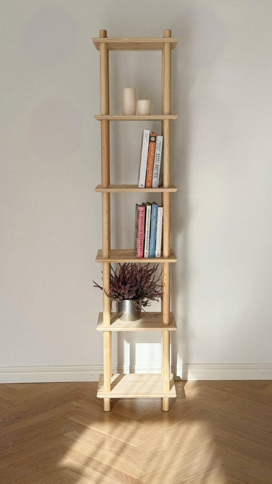 Full-height lifestyle shot of the raw pine Air Shelf narrow six-tiered unit, holding stacks of colorful books and a silver pot with purple heather, placed against a white wall and light wood floor.