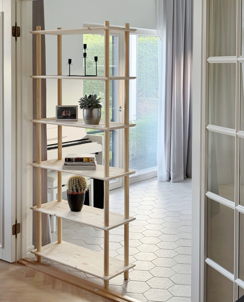 Full-height lifestyle shot of the tall, raw pine Air Shelf six-tiered unit, holding candles, books, and plants, positioned to act as a room divider between a living space and a doorway.