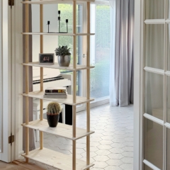 Full-height lifestyle shot of the tall, raw pine Air Shelf six-tiered unit, holding candles, books, and plants, positioned to act as a room divider between a living space and a doorway.