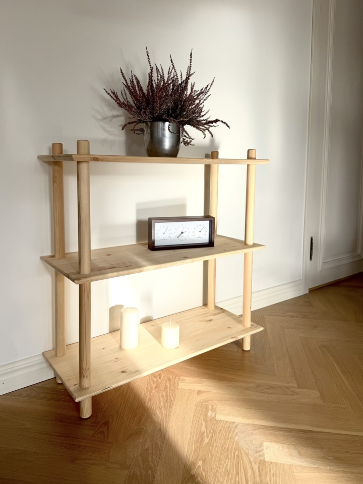 Lifestyle shot of the wide raw pine Air Shelf three-tiered unit, styled with a potted heather plant on top, a wooden clock on the middle shelf, and two white pillar candles on the bottom shelf.