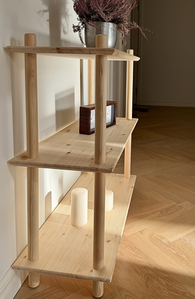 Lifestyle shot of the raw pine Air Shelf wide two-tiered modular unit, styled with a potted purple heather plant on top, a decorative wooden clock, and a book on the lower shelf, set against a white wall.