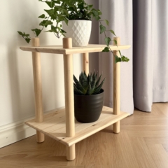 Lifestyle shot of the raw pine two-tiered modular shelf against a white wall and grey curtain, styled with two potted houseplants (succulent and ivy).