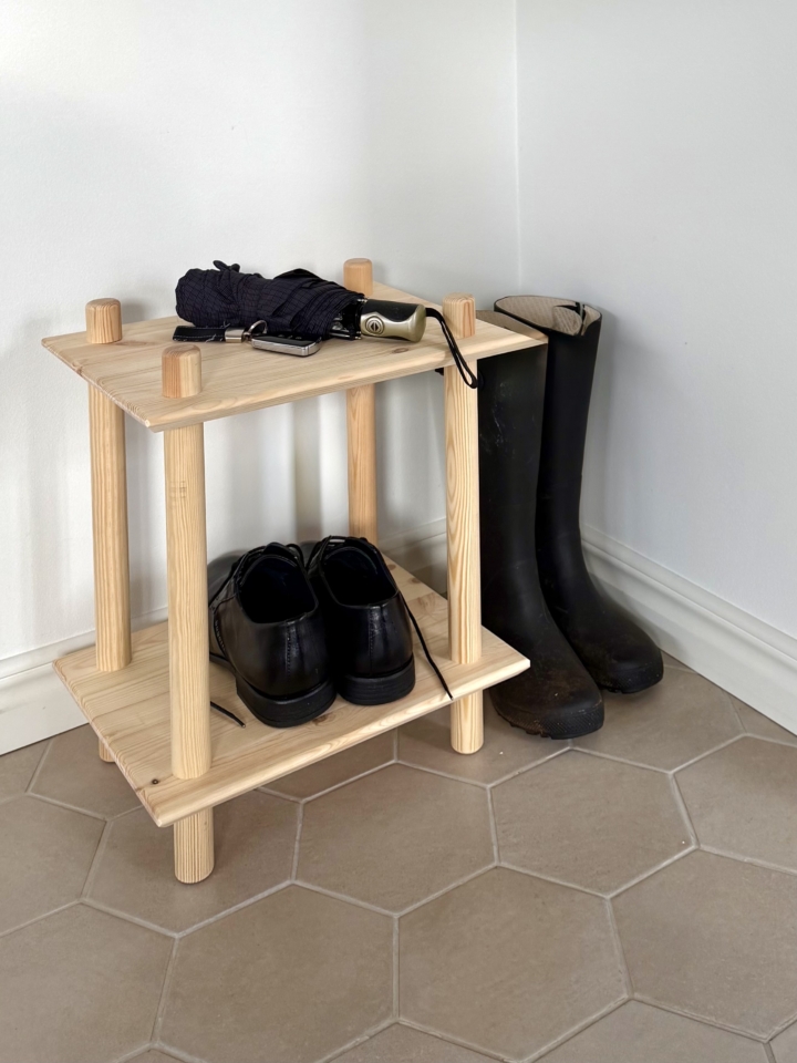 Lifestyle shot of the raw pine two-tiered modular shelf in a hallway corner, holding black shoes and an umbrella, with rain boots standing next to it.