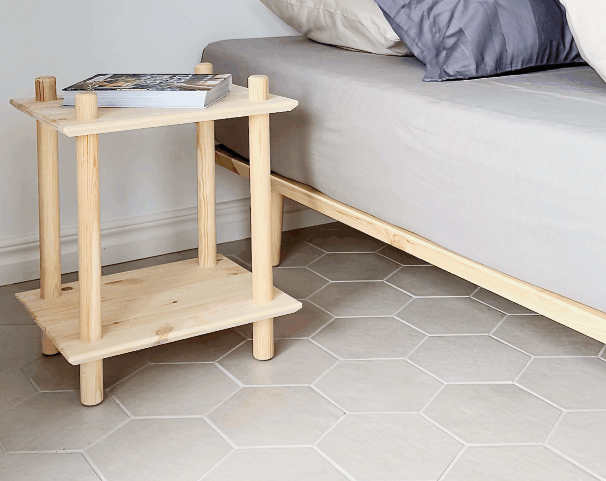 An untreated light pine Air shelf with a book on top, positioned next to an Air bed frame with neutral bedding, on a hexagonal tiled floor.