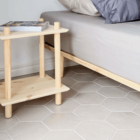 An untreated light pine Air shelf with a book on top, positioned next to an Air bed frame with neutral bedding, on a hexagonal tiled floor.