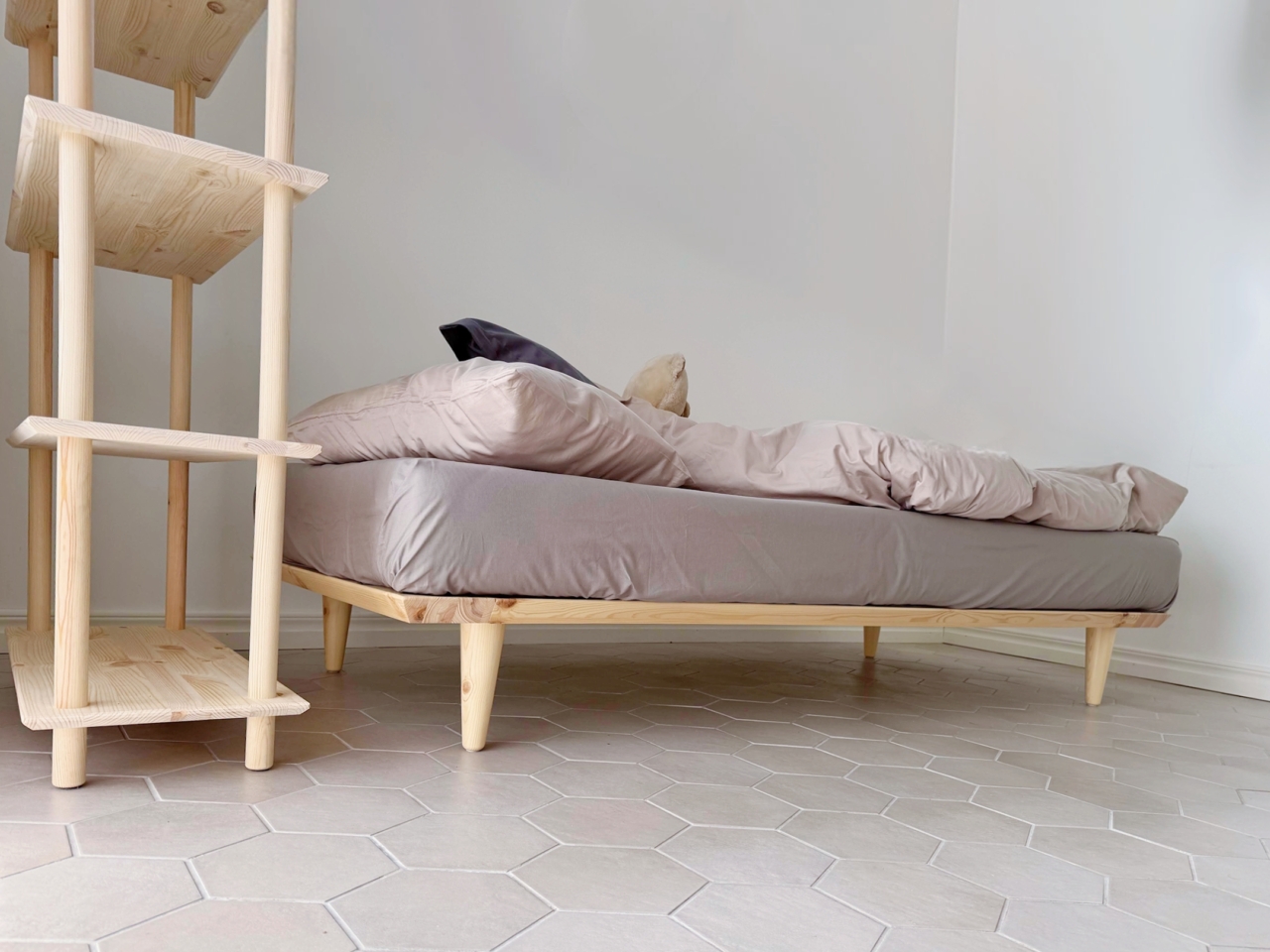 A low angle view of an untreated light pine Air bed frame 90x200 with bedding, positioned next to a matching tall Air shelf unit, on a hexagonal tiled floor.