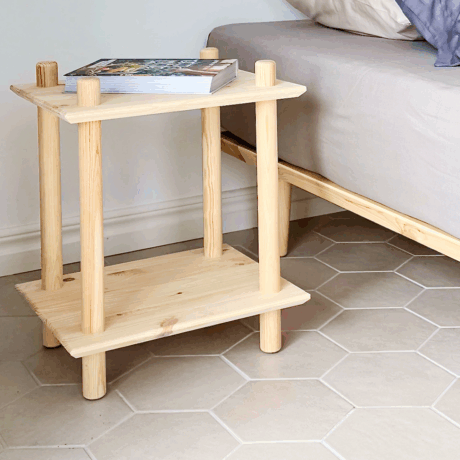 Lifestyle shot showing the raw pine Rohe two-tiered shelf functioning as a bedside table next to a bed with pillows, holding a small stack of books.