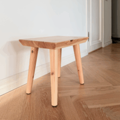 An untreated light pine Air stool stands on a herringbone parquet floor in a bright hallway, with a white wall and a closed door in the background.