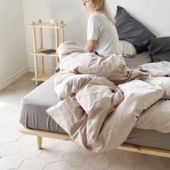 A woman sits on an untreated light pine Air Bed Frame 180x200 cm, looking out the window, with a narrow Air Shelf next to the bed and neutral bedding.