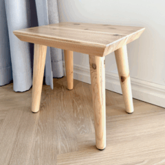 Close-up of an untreated light pine Air stool on a light wooden floor, showing the natural wood grain and subtle knots, with a curtain in the background.