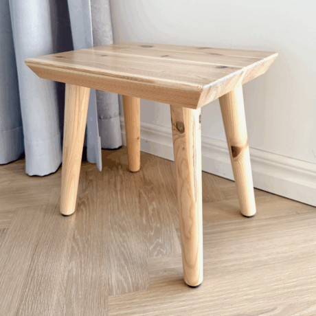 Close-up of an untreated light pine Air stool on a light wooden floor, showing the natural wood grain and subtle knots, with a curtain in the background.