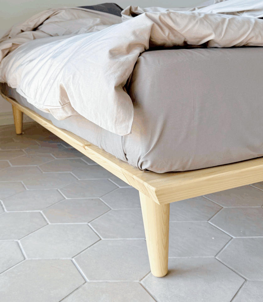 Close-up of the tapered wooden leg and corner frame of an untreated light pine Air bed frame 180x200 cm, with bedding, on a hexagonal tiled floor.