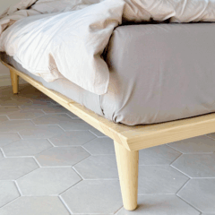 Close-up of the tapered wooden leg and corner frame of an untreated light pine Air bed frame 180x200 cm, with bedding, on a hexagonal tiled floor.