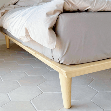 Close-up of the tapered wooden leg and corner frame of an untreated light pine Air bed frame 180x200 cm, with bedding, on a hexagonal tiled floor.