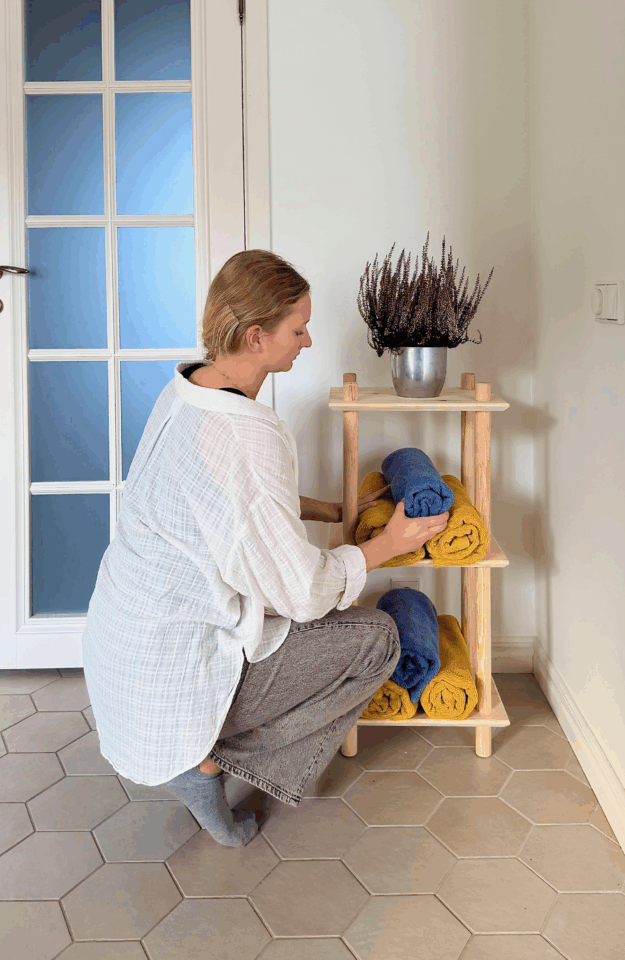 A woman in a white blouse is crouching, carefully placing a blue rolled towel onto the raw pine Air Shelf narrow three-tiered modular unit, which also holds yellow towels and a potted plant.