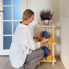 A woman in a white blouse is crouching, carefully placing a blue rolled towel onto the raw pine Air Shelf narrow three-tiered modular unit, which also holds yellow towels and a potted plant.