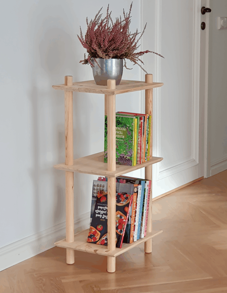 Lifestyle shot of the raw pine Air Shelf narrow three-tiered unit, styled with a silver pot of purple heather on top and several colorful books and cookbooks arranged on the two lower shelves.