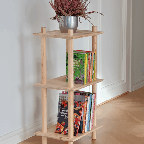 Lifestyle shot of the raw pine Air Shelf narrow three-tiered unit, styled with a silver pot of purple heather on top and several colorful books and cookbooks arranged on the two lower shelves.