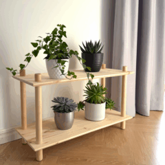 Lifestyle shot of the raw pine Air Shelf wide two-tiered modular unit, filled with four potted houseplants (ivy, succulents) against a white wall and grey curtain, on a light wooden floor.