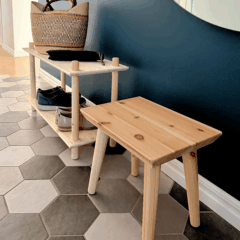 Lifestyle shot featuring the raw pine Air Shelf two-tiered unit holding a woven basket and shoes, placed next to the Air Stool, against a dark blue wall with a round mirror.