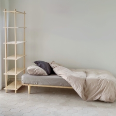 An untreated light pine Air bed frame 90x200 with neutral bedding is placed next to a tall modular Air shelf unit in a bright, minimalist room.