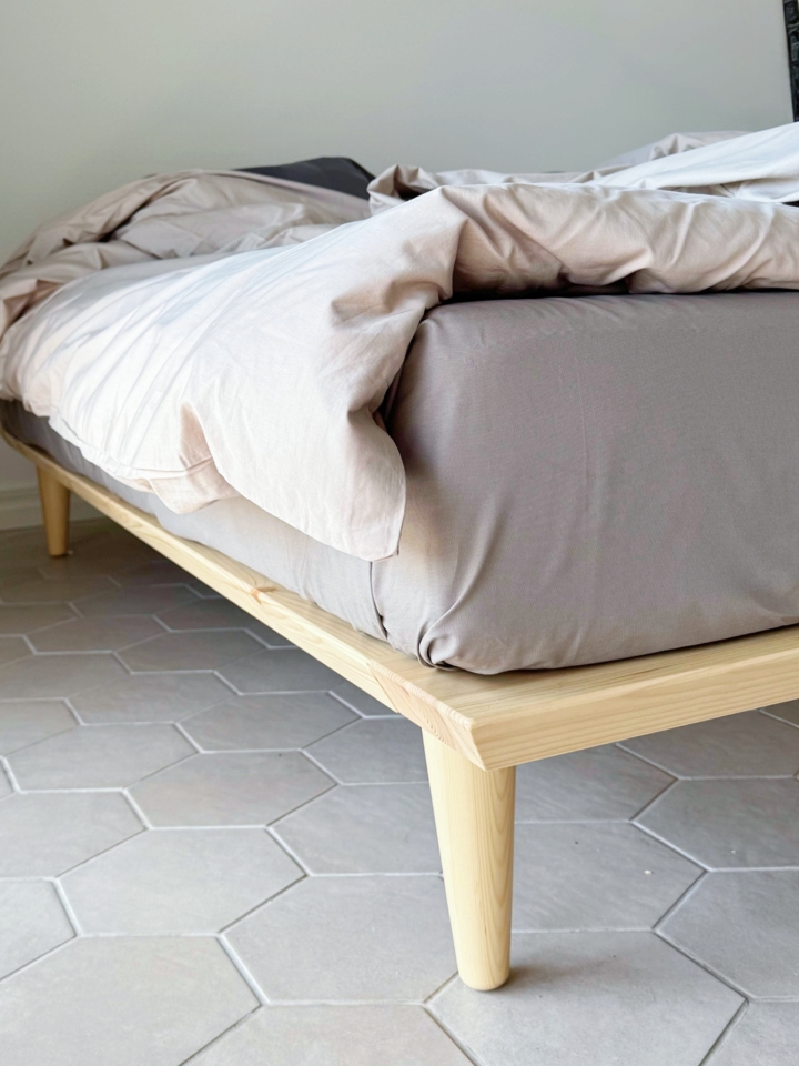Close-up of the tapered wooden leg and corner frame of an untreated light pine Air bed frame 160x200 cm, with bedding, on a hexagonal tiled floor.