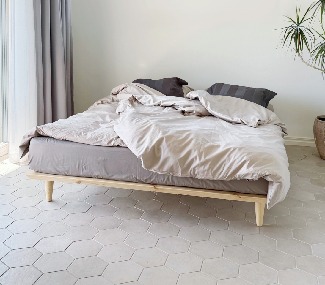 An untreated light pine Air bed frame 140x200 cm with grey and light beige bedding, placed on a light hexagonal tiled floor in a minimalist bedroom.